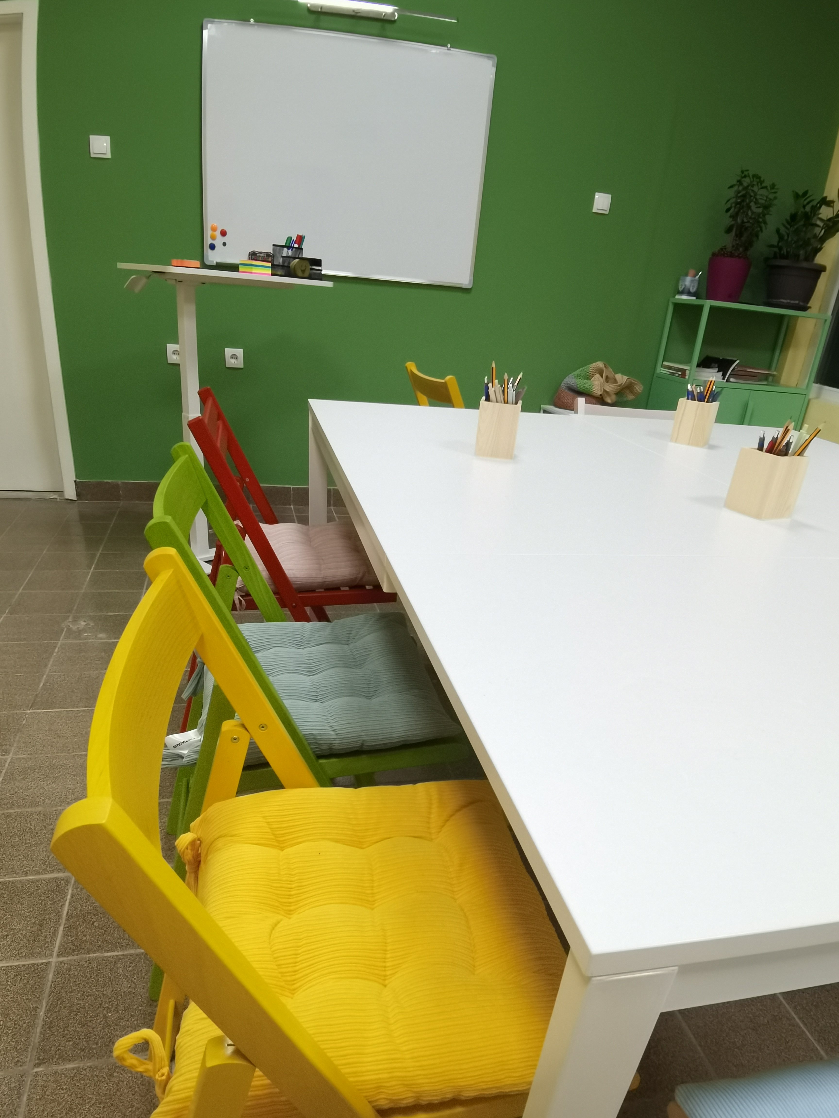 Učionica sa belim stolom, obojenim stolicama i tablom na zelenoj zidu